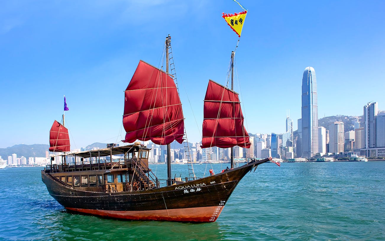 AquaLuna junk boat sailing in Hong Kong Harbor with city skyline in the background.