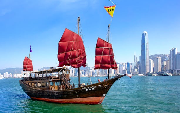 AquaLuna junk boat sailing in Hong Kong Harbor with city skyline in the background.