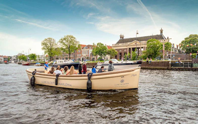 Open boat with tourists cruising Haarlem canal, passing historic buildings and trees.