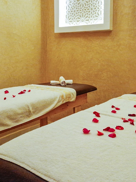 Spa treatment room with two massage tables, rose petals, and rolled towels.