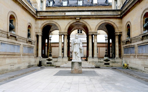 Statue in the courtyard of Collège de France, Paris, framed by arches and classical architecture.