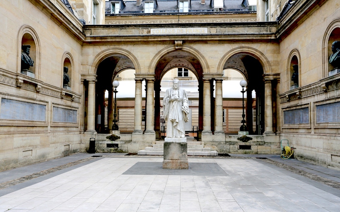 Statue in the courtyard of Collège de France, Paris, framed by arches and classical architecture.