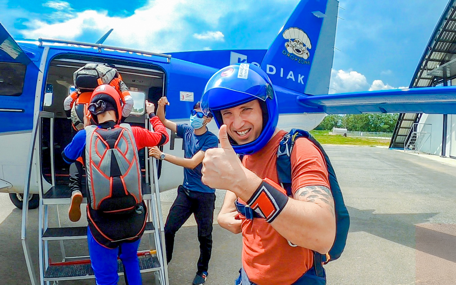 Tourists boarding a skydive plane, one giving a thumbs up, at a dropzone.
