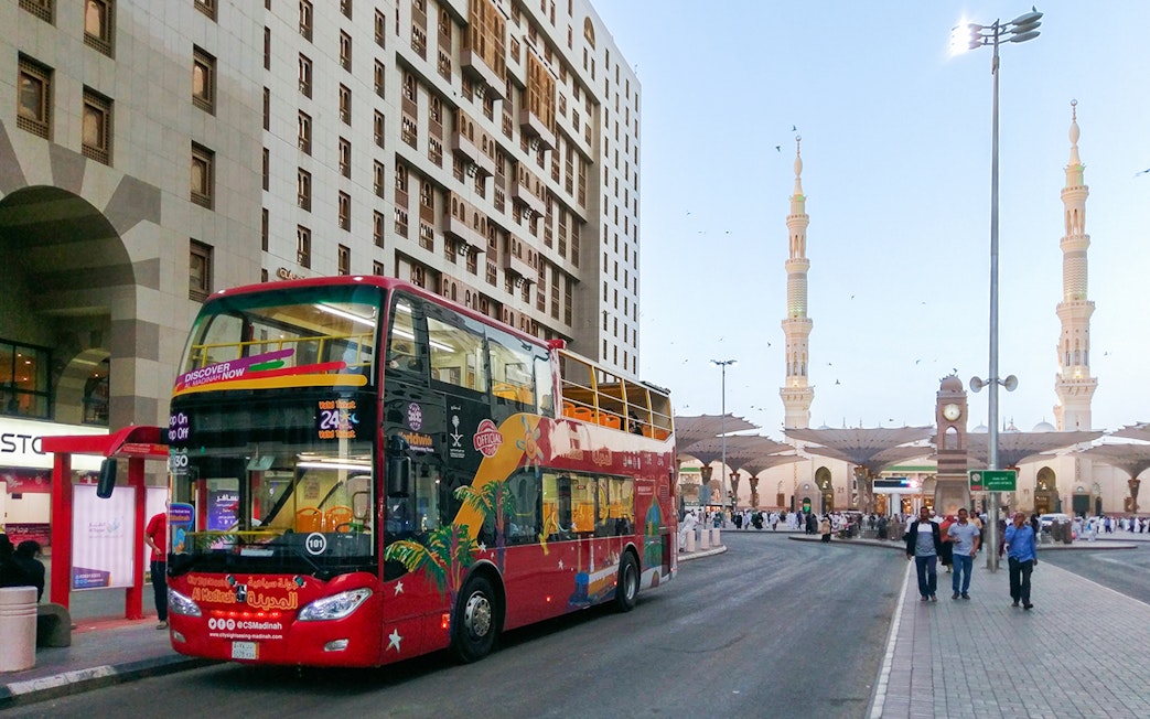 Red double-decker bus on Al Madinah Hop-On Hop-Off Tour near mosque and city buildings.