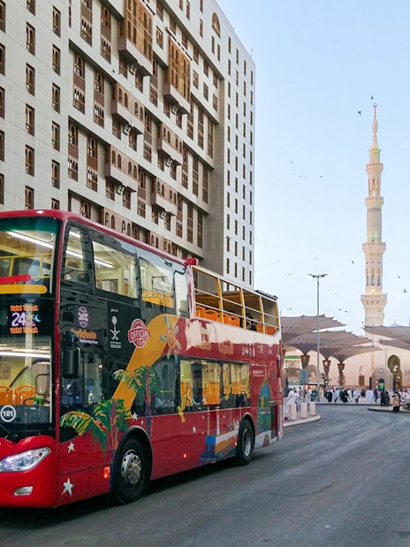Red double-decker bus on Al Madinah Hop-On Hop-Off Tour near mosque and city buildings.