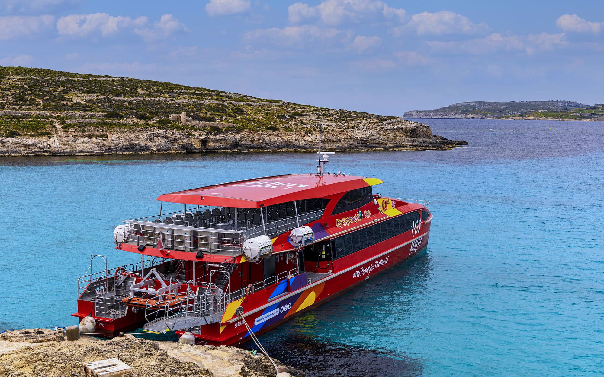 Catamaran docked in clear blue waters of Malta with rocky coastline in the background.