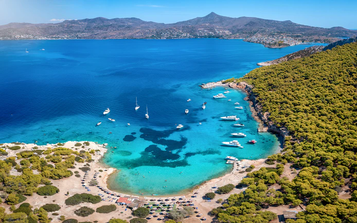 Boats anchored in a turquoise bay near Athens, surrounded by lush greenery and distant mountains.