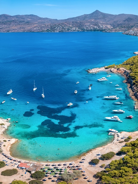 Boats anchored in a turquoise bay near Athens, surrounded by lush greenery and distant mountains.