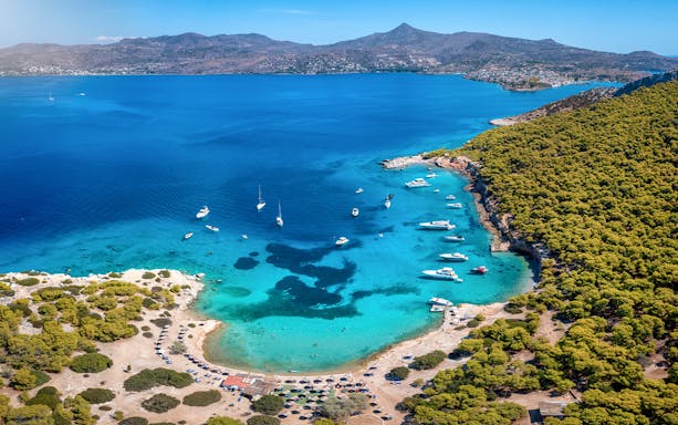 Boats anchored in a turquoise bay near Athens, surrounded by lush greenery and distant mountains.