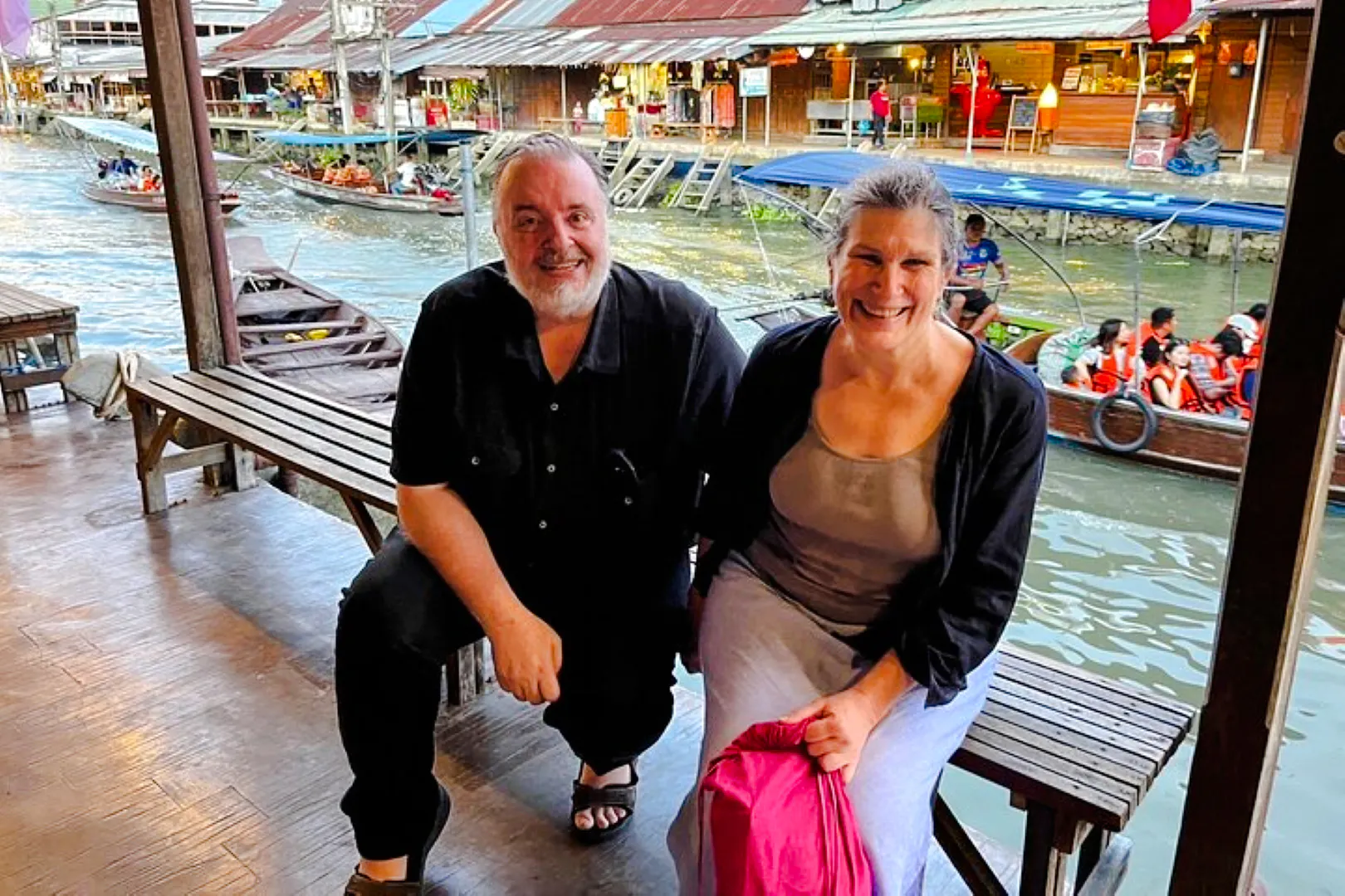 Couple sitting by Amphawa Floating Market canal with boats and shops in background, Pattaya excursion.