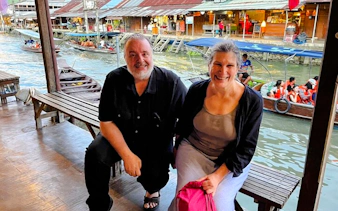Couple sitting by Amphawa Floating Market canal with boats and shops in background, Pattaya excursion.