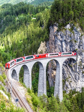 Red train crossing the Landwasser Viaduct in Switzerland, surrounded by lush green forest.