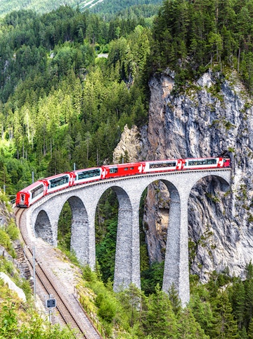 Red train crossing the Landwasser Viaduct in Switzerland, surrounded by lush green forest.
