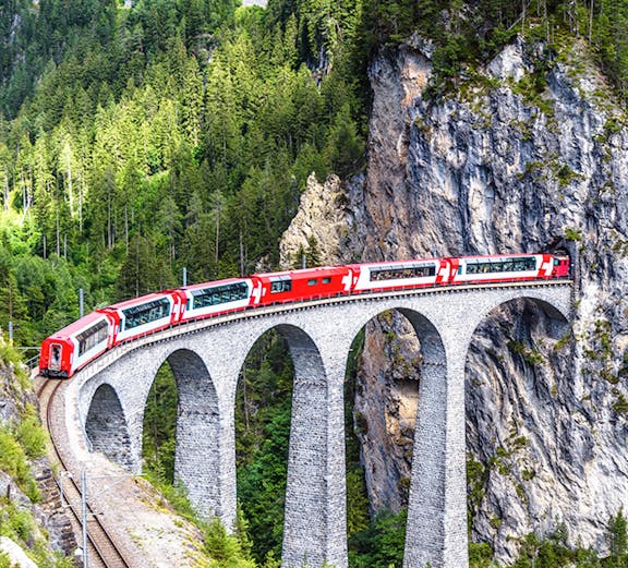 Red train crossing the Landwasser Viaduct in Switzerland, surrounded by lush green forest.