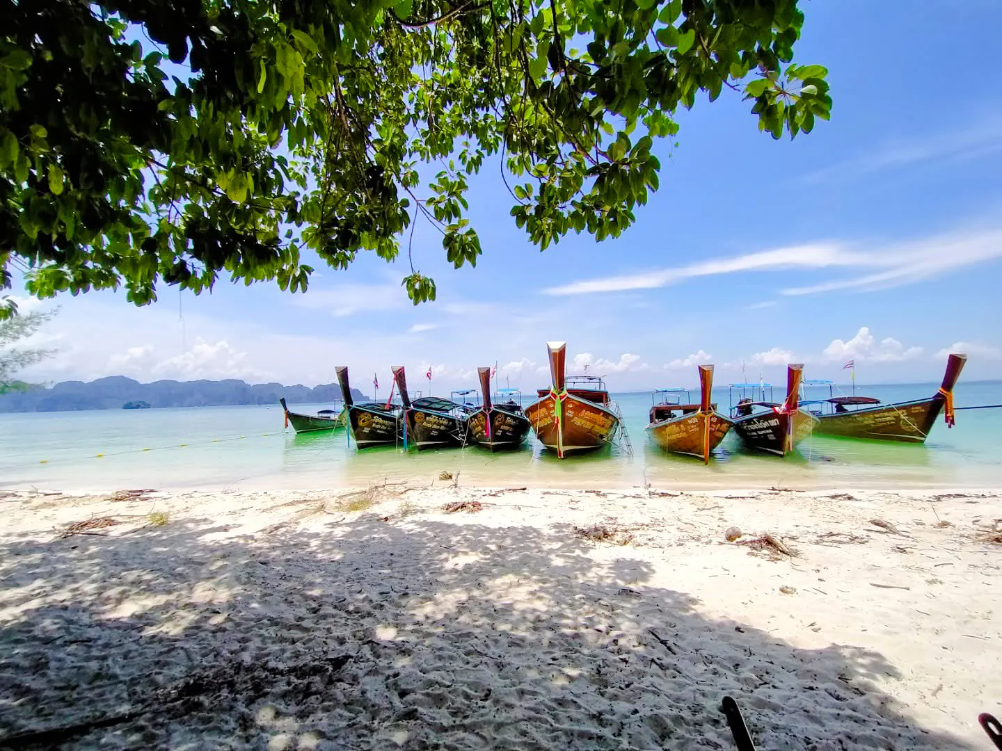 Longtail boats anchored on a sandy beach in Krabi, Thailand, with clear blue water and distant islands.