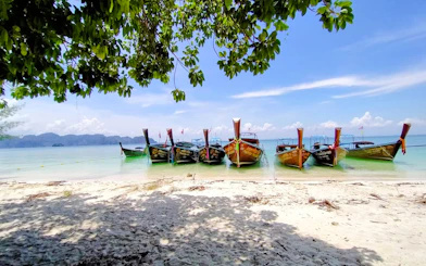 Longtail boats anchored on a sandy beach in Krabi, Thailand, with clear blue water and distant islands.