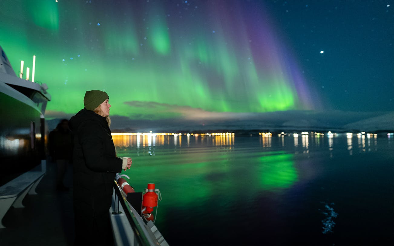 Woman on cruise viewing deck watching Northern Lights over water.