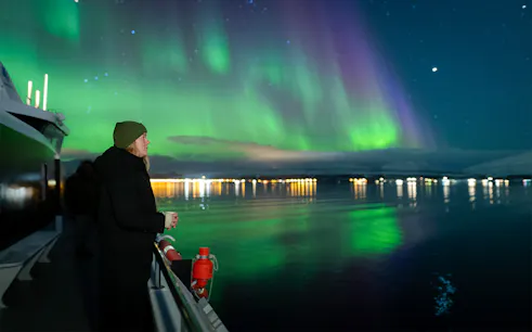 Woman on cruise viewing deck watching Northern Lights over water.