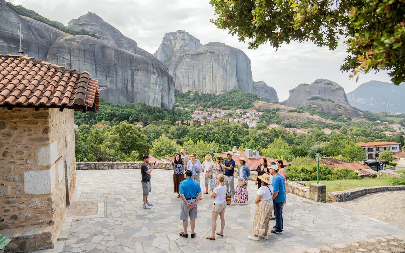Local guide with group near Meteora monastery, Greece, gesturing towards rock formations.