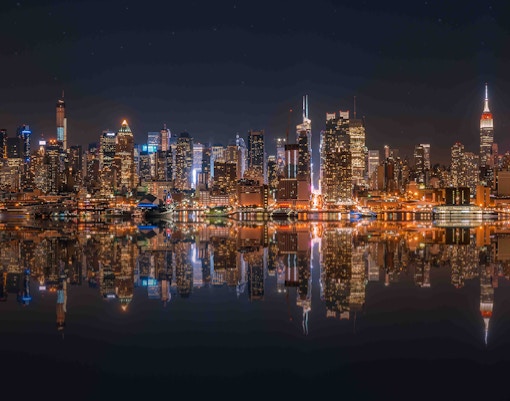 New York City skyline at night reflected on the Hudson River.