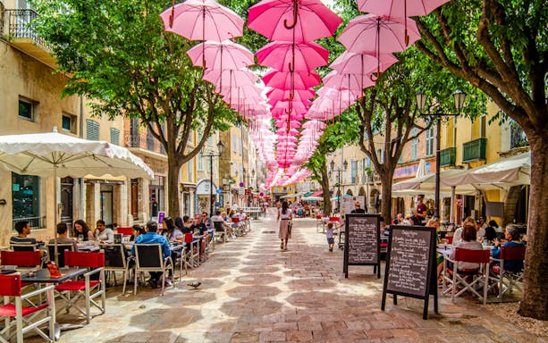Street in Provence with pink umbrellas overhead and outdoor cafes.