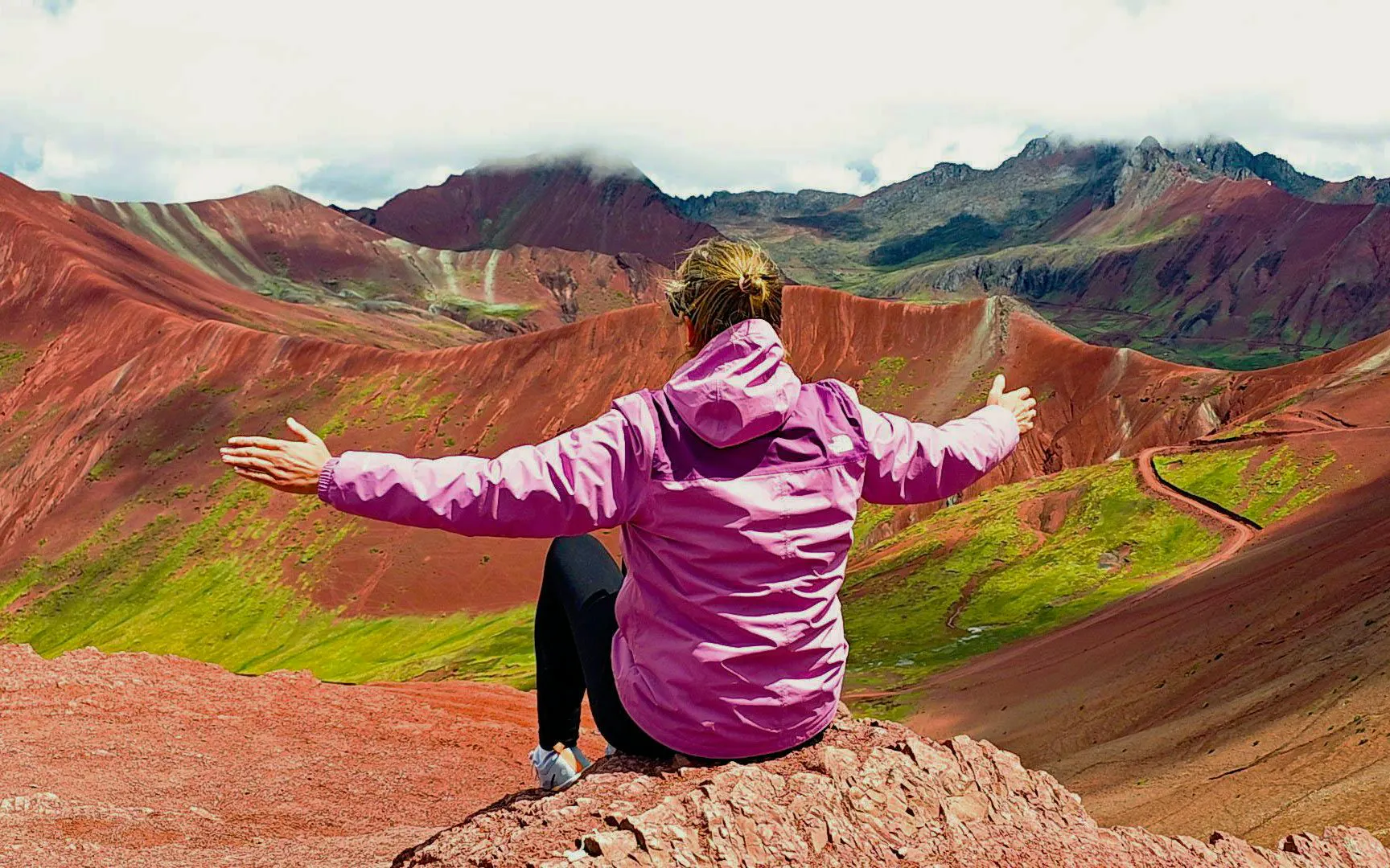 Person sitting on Rainbow Mountain, Peru, overlooking colorful Red Valley landscape.