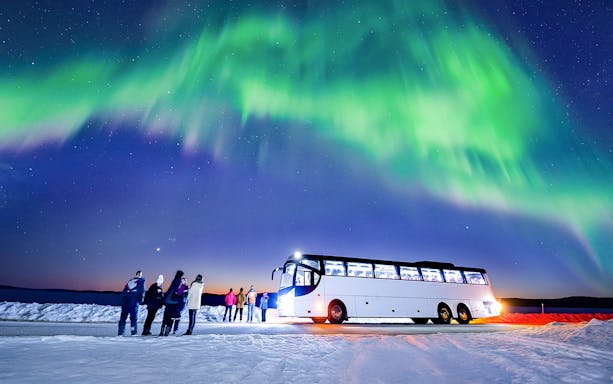 Tourists stand near a bus under the Northern Lights in a snowy landscape.