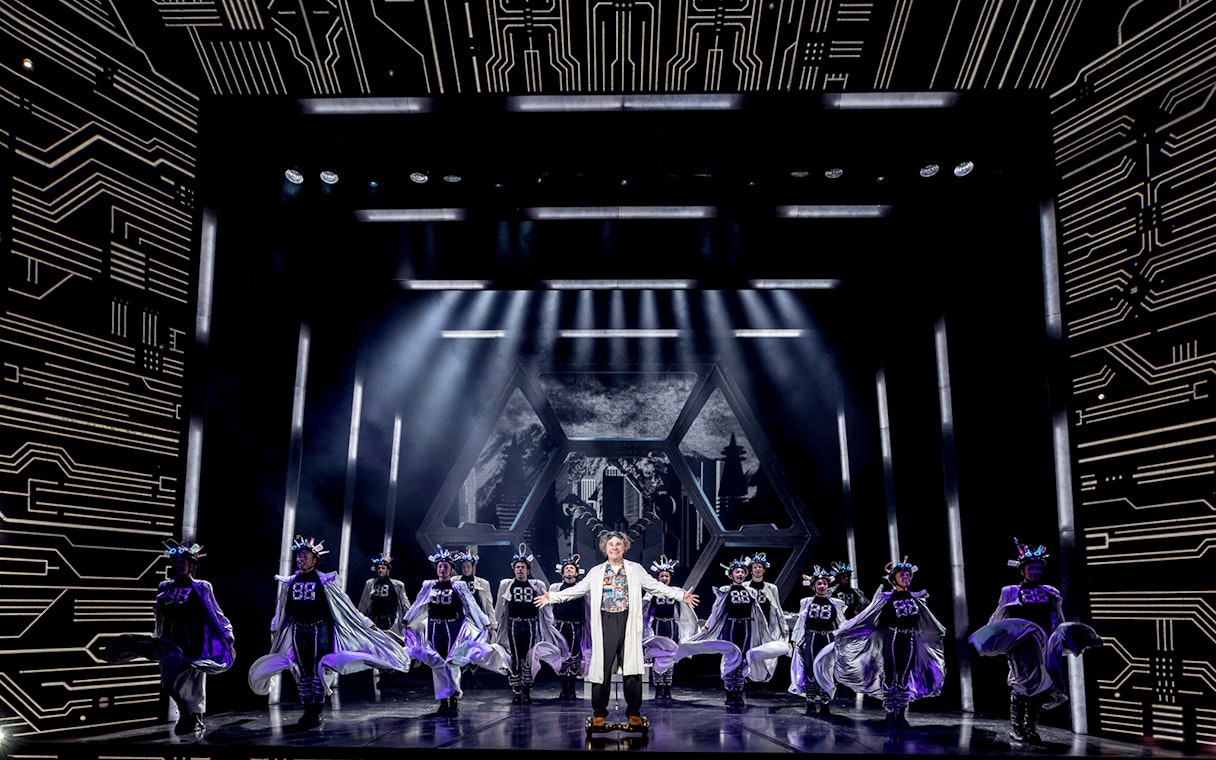 Stage performance of "Back to the Future" with actors in futuristic costumes and dynamic lighting.