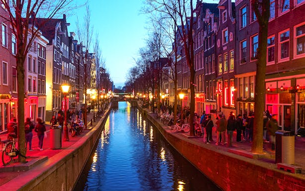 Canal view with red-lit buildings and people walking in Amsterdam's Red Light District.