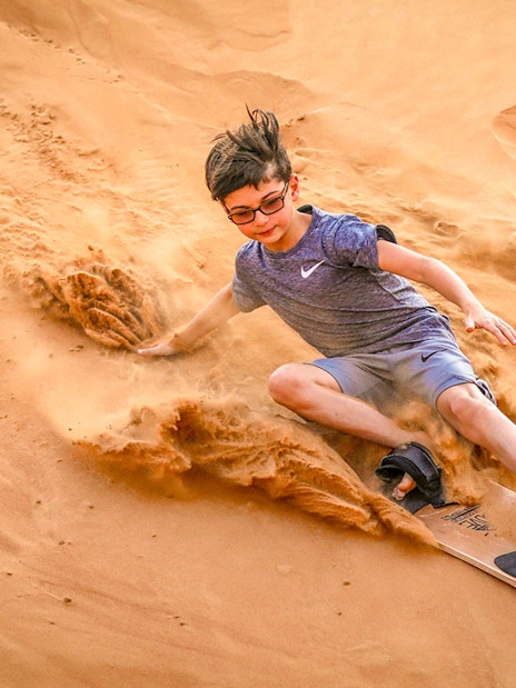 Young person sandboarding down a dune during a desert safari in Dubai.