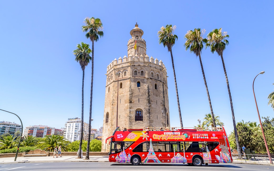 Seville sightseeing bus in front of Torre del Oro with palm trees.