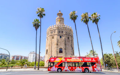 Seville sightseeing bus in front of Torre del Oro with palm trees.
