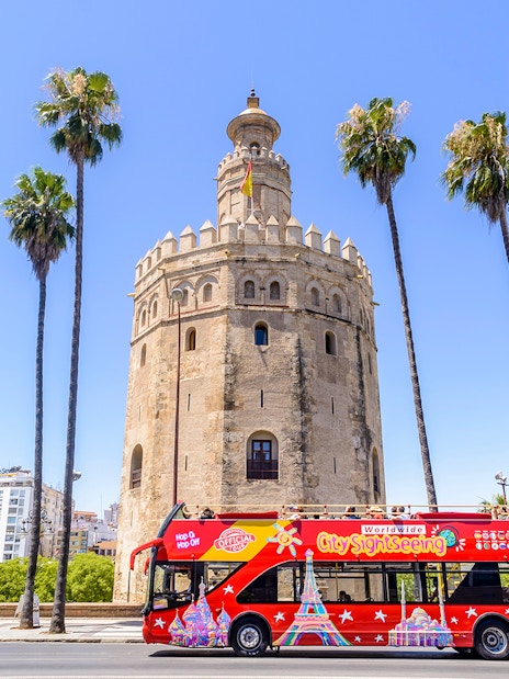 Seville sightseeing bus in front of Torre del Oro with palm trees.