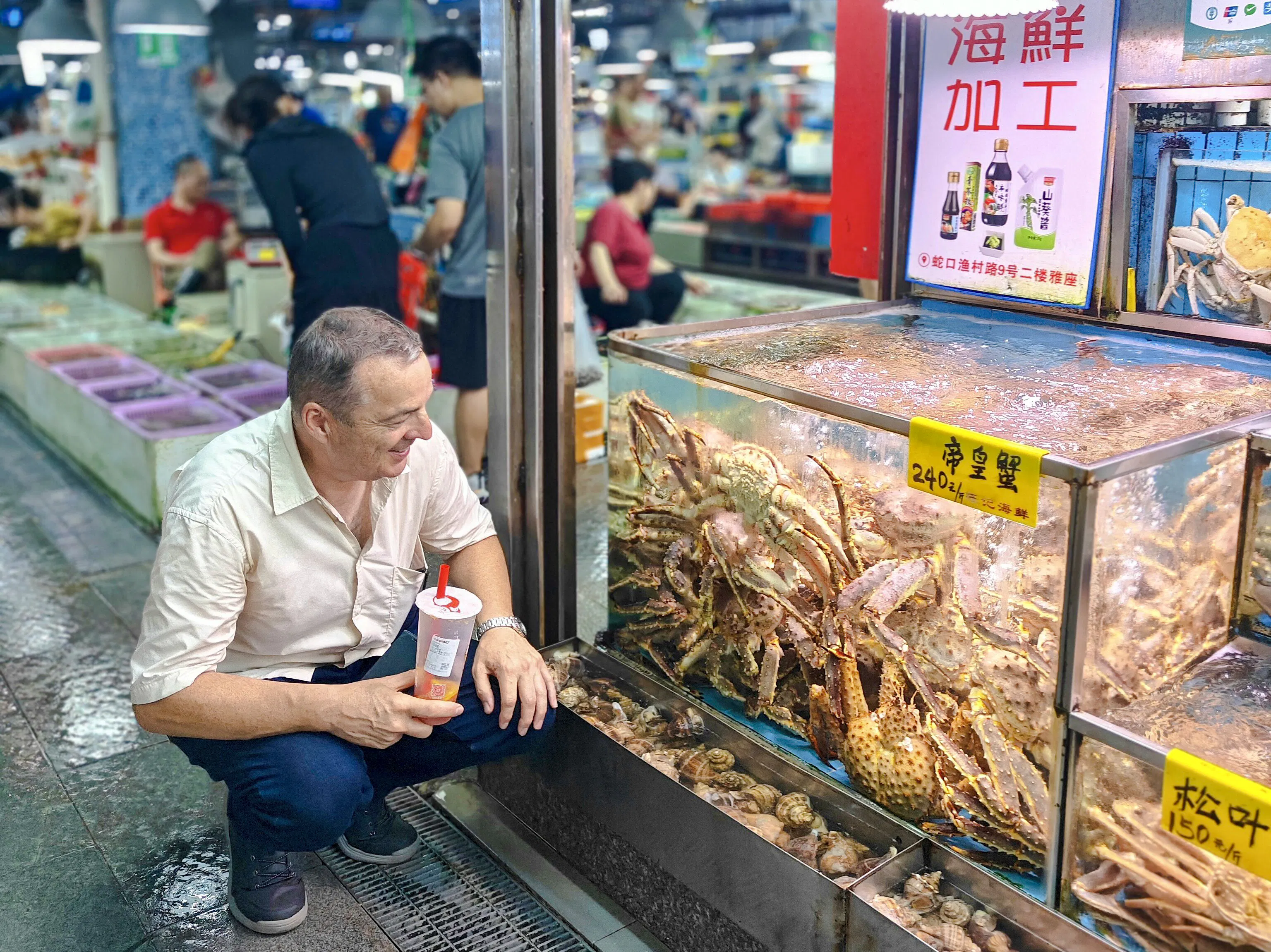 Man observing seafood tank in Shenzhen market, holding a drink, surrounded by local vendors and shoppers.