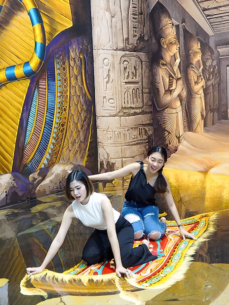 Two visitors on a 3D magic carpet illusion at Art in Paradise Pattaya, with Egyptian-themed murals.