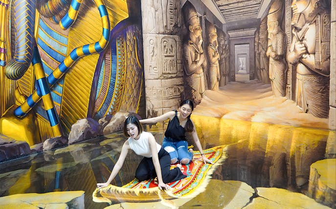 Two visitors on a 3D magic carpet illusion at Art in Paradise Pattaya, with Egyptian-themed murals.