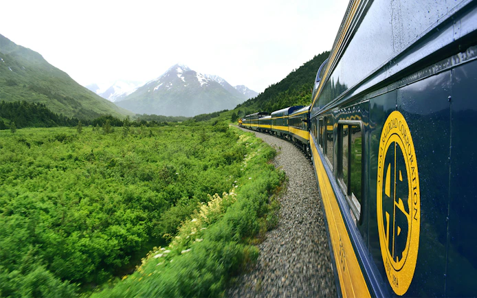 Alaska Railroad train traveling through lush green landscape with mountains in the background.