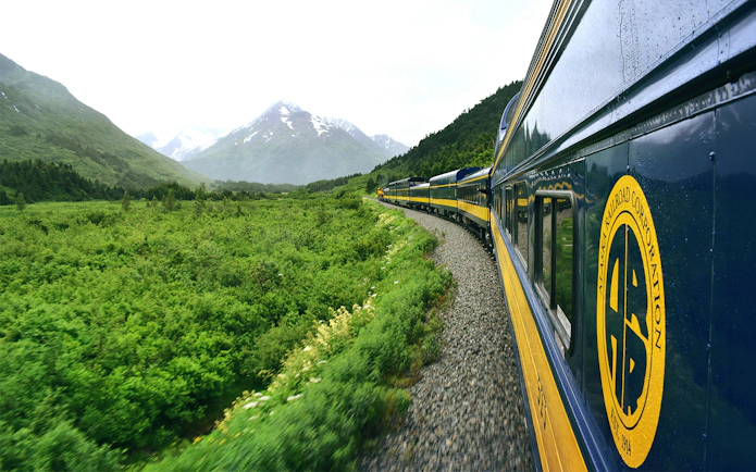 Alaska Railroad train traveling through lush green landscape with mountains in the background.