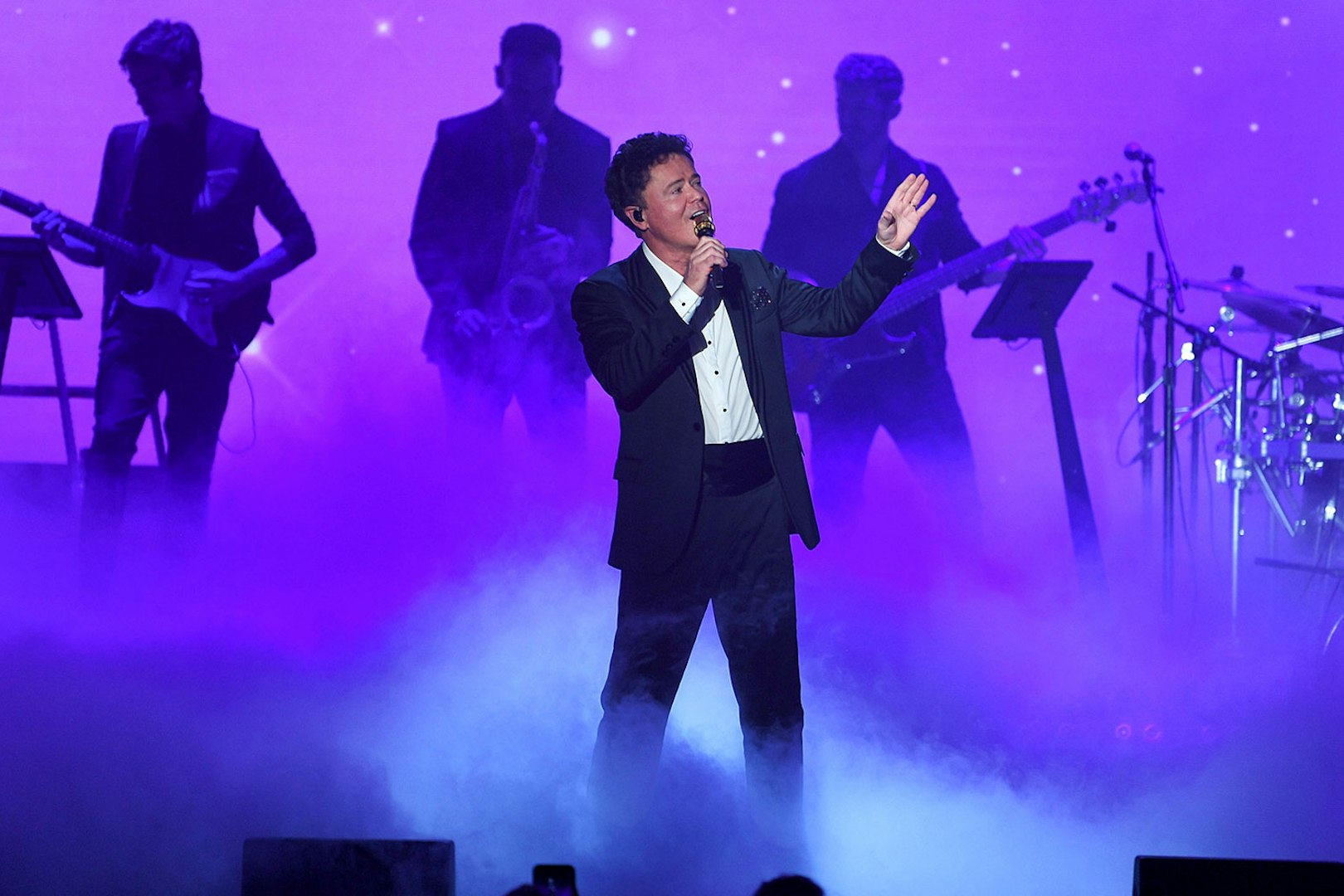 Singer performing on stage with a band in a purple-lit setting.