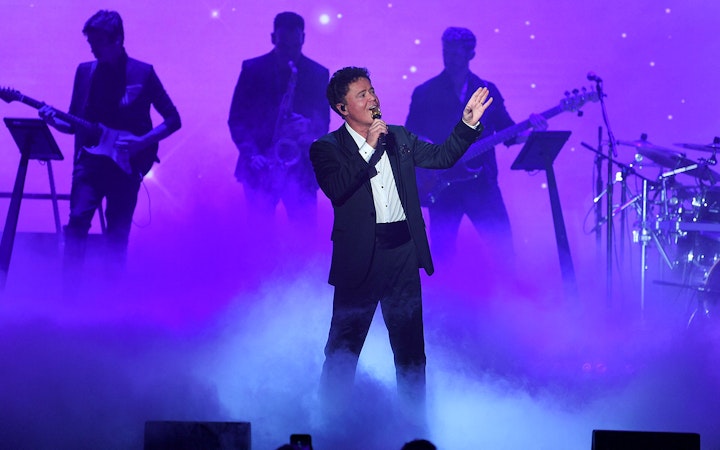 Singer performing on stage with a band in a purple-lit setting.