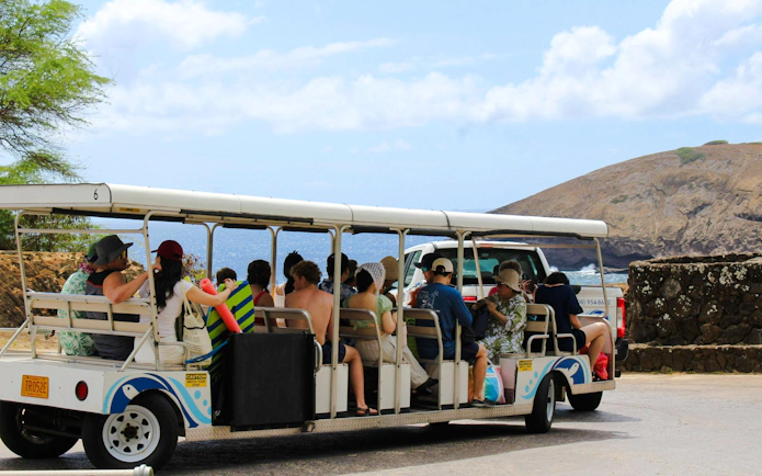 Tourists on a tram at Hanauma Bay Nature Preserve with ocean and cliffs in the background.