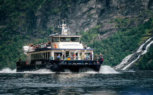 Cruise boat on Geiranger Fjord with passengers, surrounded by cliffs and waterfall, Geiranger.