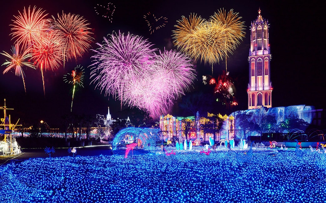 Fireworks display over illuminated Huis Ten Bosch theme park at night.