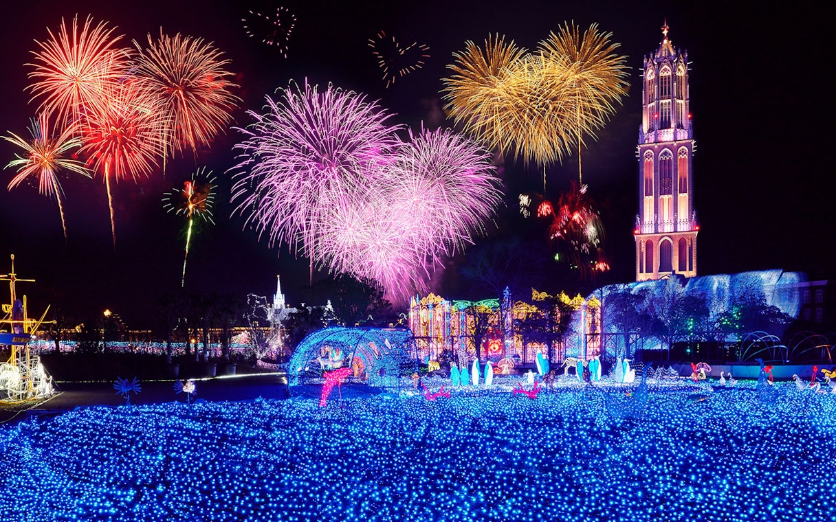 Fireworks display over illuminated Huis Ten Bosch theme park at night.