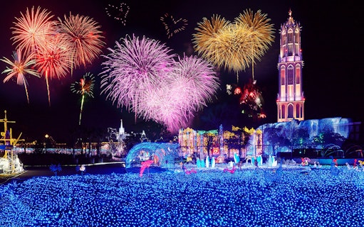 Fireworks display over illuminated Huis Ten Bosch theme park at night.