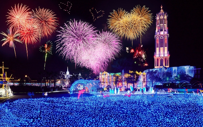 Fireworks display over illuminated Huis Ten Bosch theme park at night.
