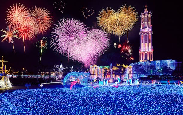 Fireworks display over illuminated Huis Ten Bosch theme park at night.