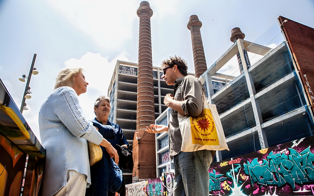 Tour guide discussing Barcelona's Raval history with visitors near graffiti-covered walls and industrial chimneys.