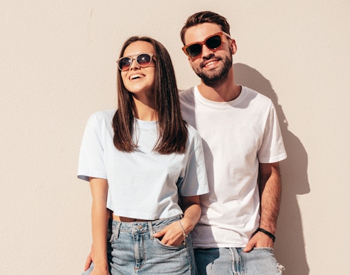 Couple in casual clothes and sunglasses standing against a beige wall.