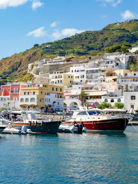Boats docked at a colorful harbor in Ischia with hillside houses in the background.